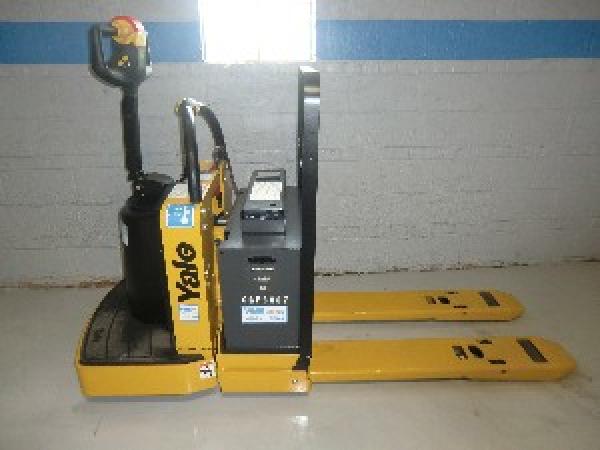 A stationary black and yellow Yale pallet jack sits in front of a grey warehouse wall with blue trimming.