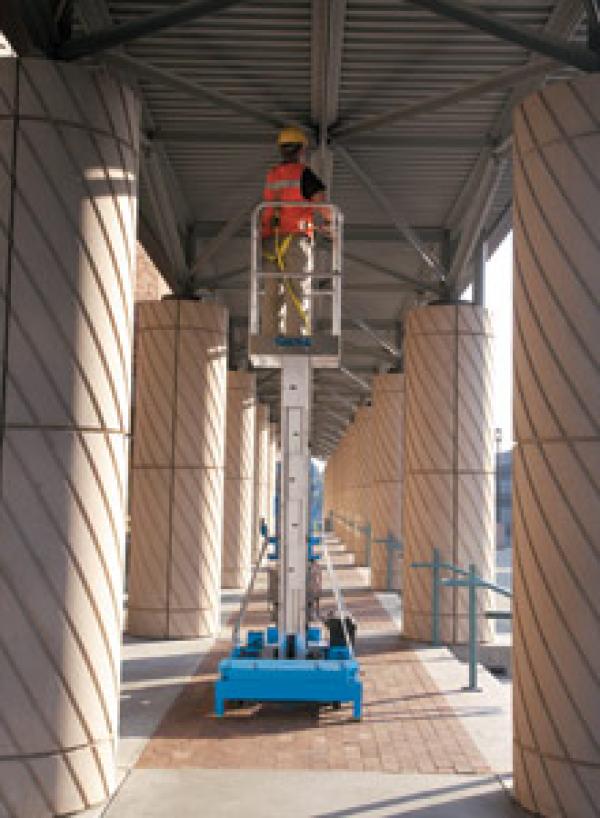 A man in a yellow hard hat and an orange safety vest is fixing the ceiling while extended on a piece of blue lift machinery.