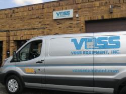 title A silver van is parked next to a brick building with the material handling brand, Voss Equipment's image on the van.