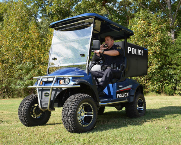 A blue and black electric police cart is parked on a patch of grass in front of a tree line while an officer sits inside of it.