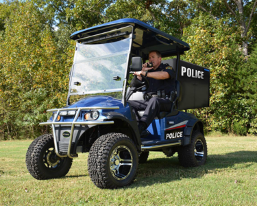 A blue and black electric police cart is parked on a patch of grass in front of a tree line with an officer inside of it.