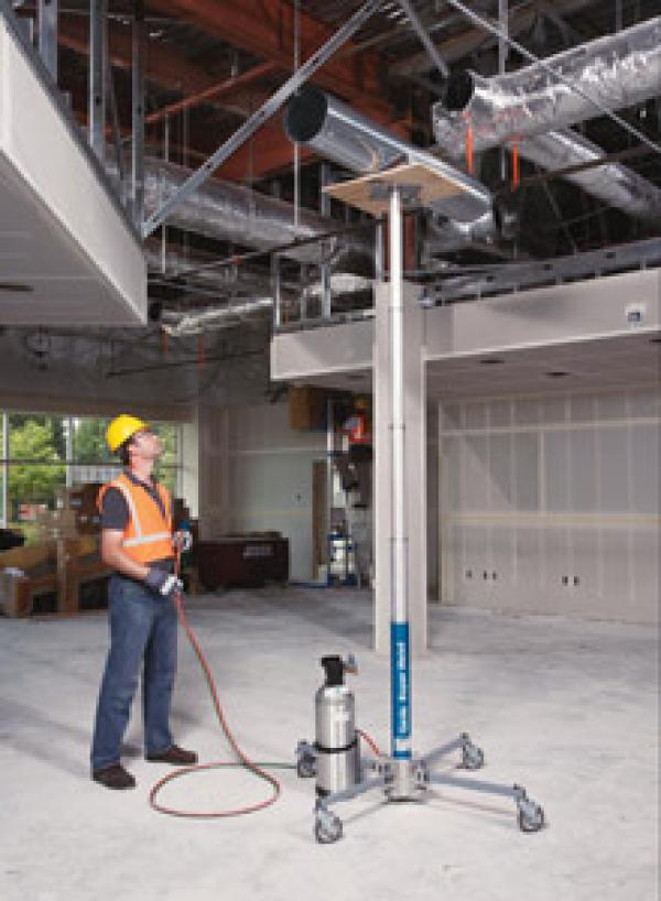 A man in a yellow hard hat and orange safety vest is operating a hydraulic jack lift to lift ventilation to the ceiling.